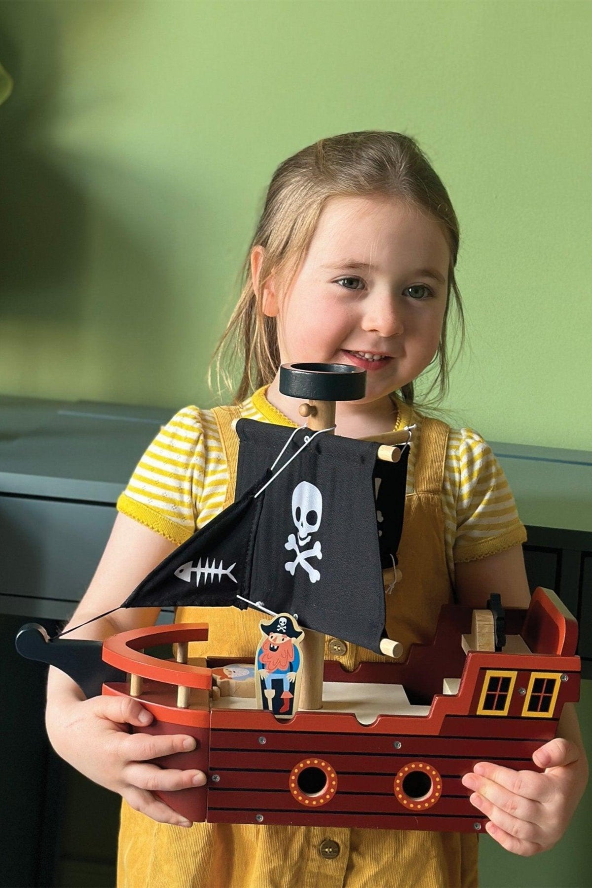 A child holds a wooden pirate ship toy featuring black sails with skull and fishbone designs.