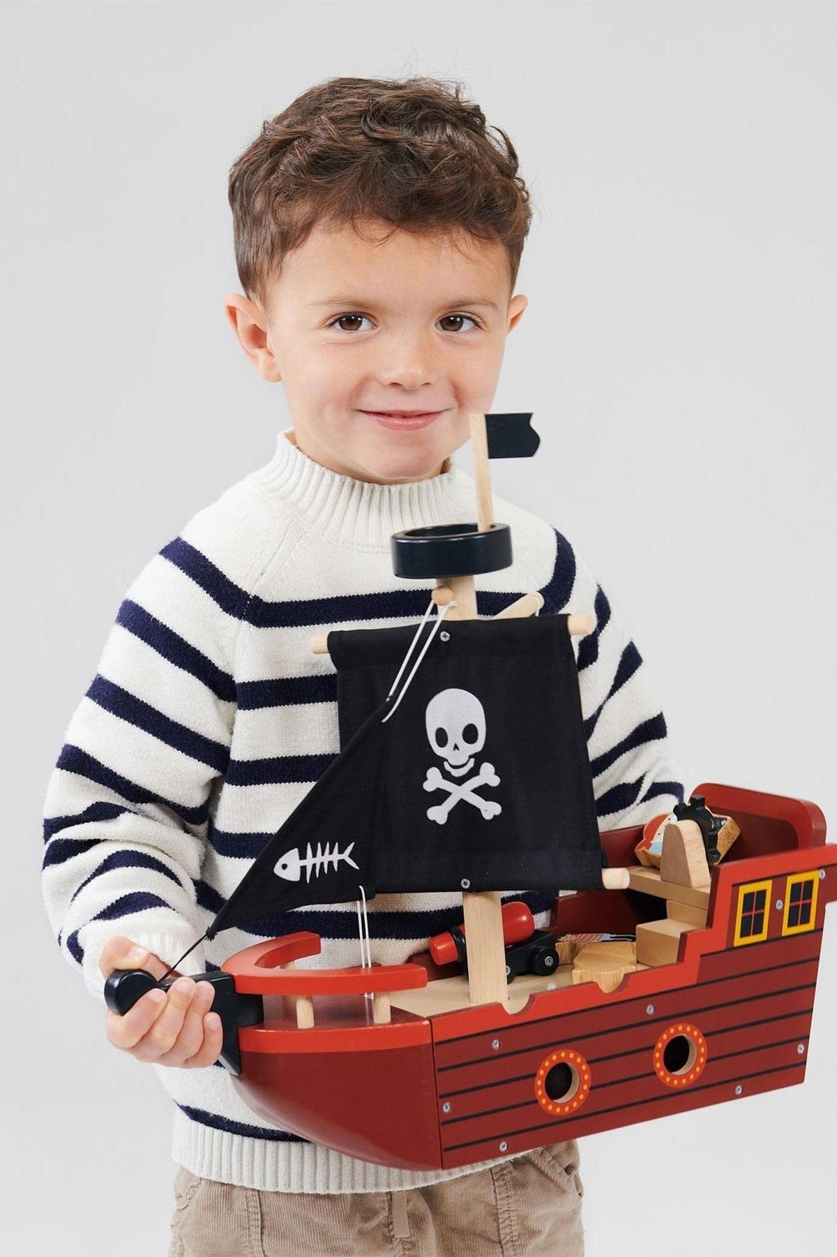 A child in a striped sweater holds a red wooden pirate ship with a black sail featuring a skull.