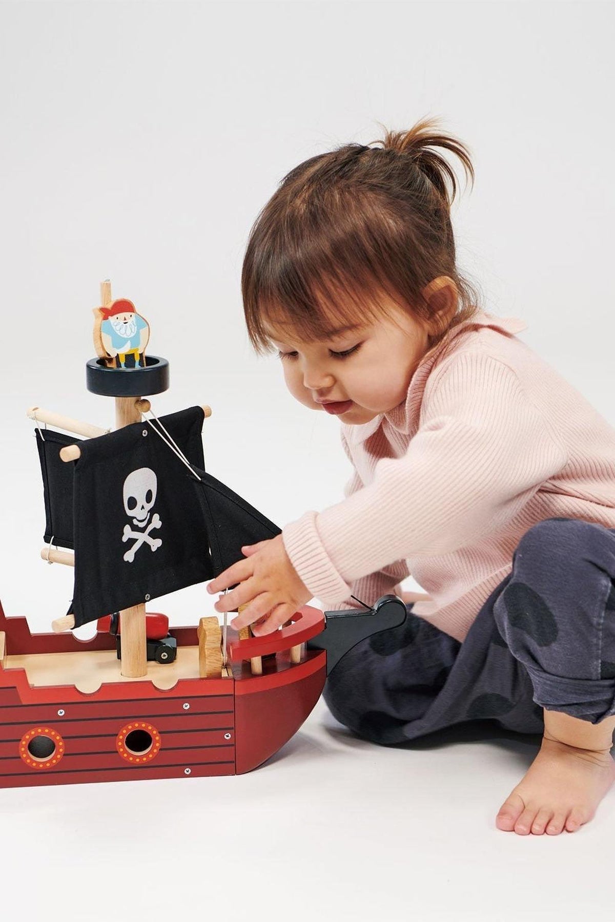 A child plays with a red wooden pirate ship featuring black sails and a skull design, sitting on the floor.