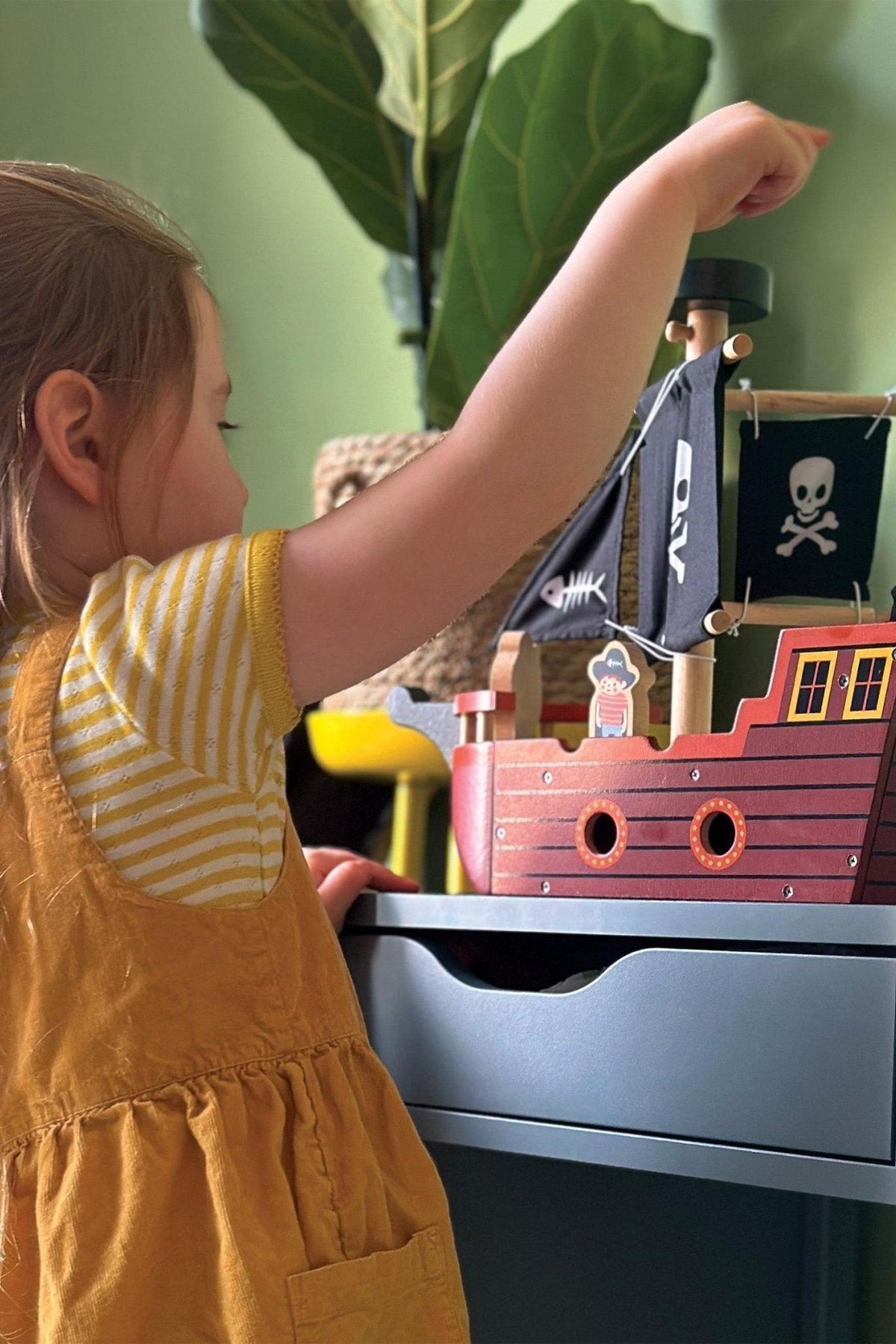 A child plays with a wooden pirate ship featuring black sails with skull and fishbone designs.