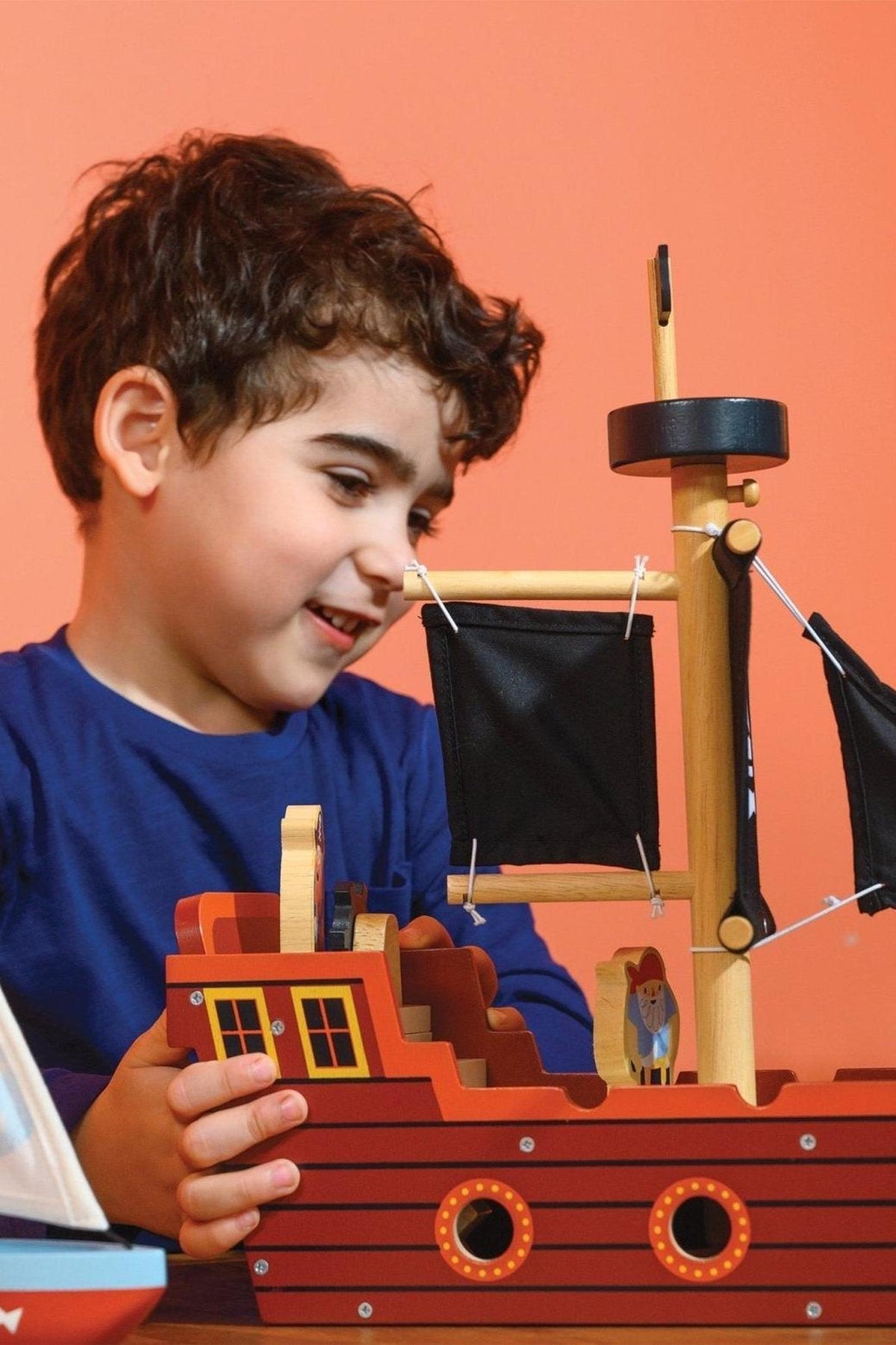 A child plays with a wooden pirate ship toy featuring black fabric sails against an orange background.