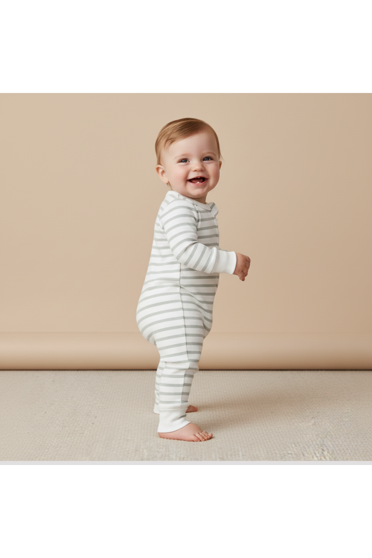 A smiling baby stands sideways, wearing a soft, organic cotton romper in fog stripes. The romper features fold-over mittens and foot cuffs, ideal for comfort and easy changing.