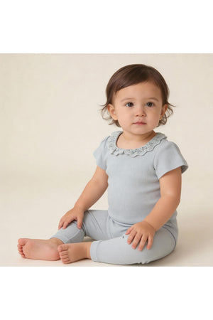 A young child sits on a light background, wearing a soft, organic cotton onesie and matching tights in a gentle fog color, featuring a delicate lace collar.