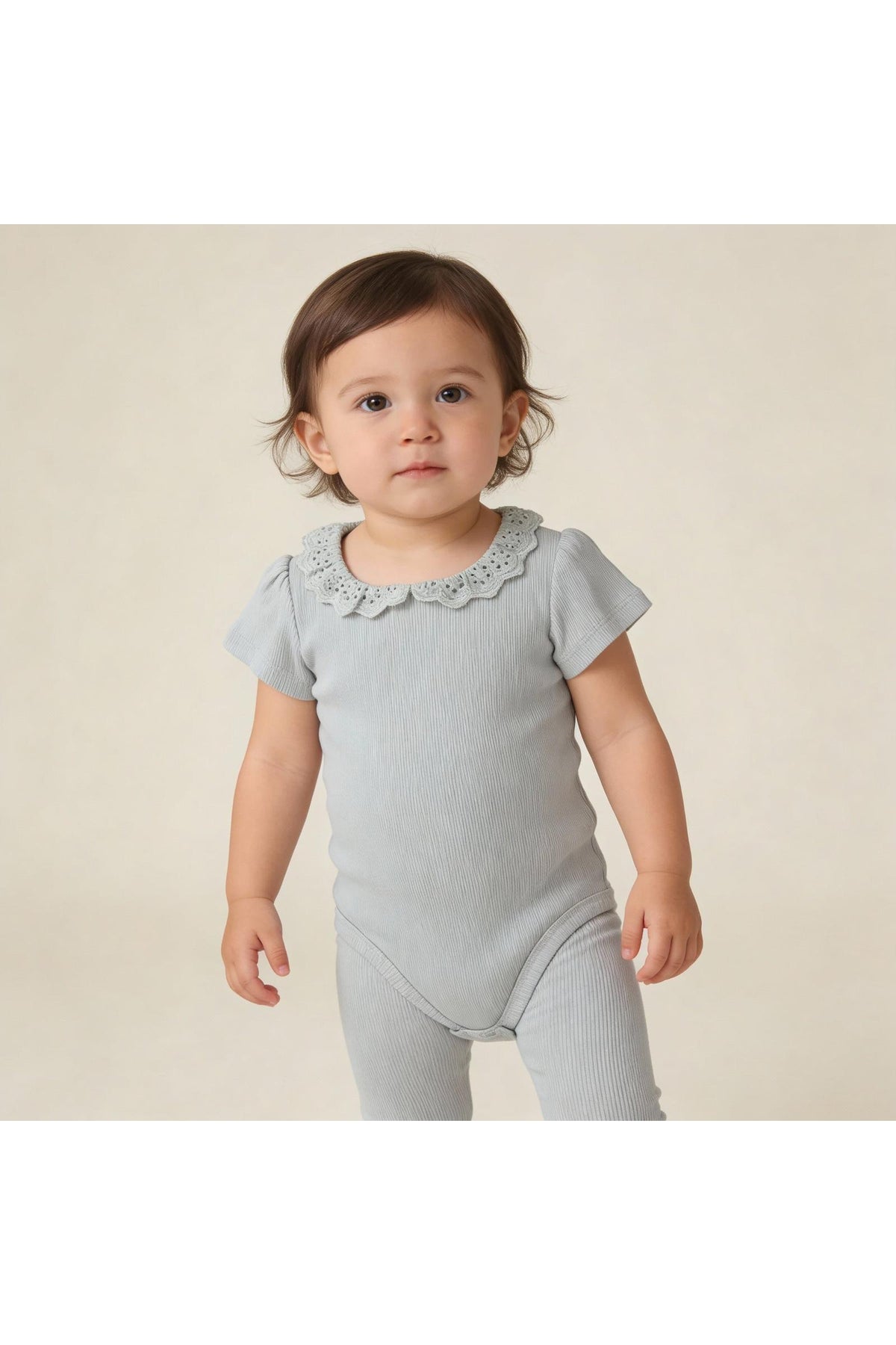 A young child stands wearing a soft, light gray organic cotton onesie with a delicate lace collar. The cozy ribbed texture is perfect for comfortable play and everyday wear.