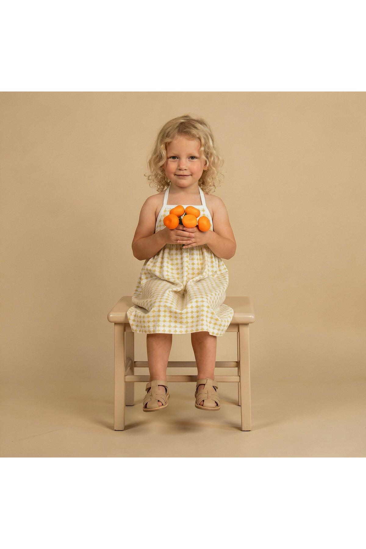 A young girl sits on a light wooden chair, wearing a yellow and white gingham tie-back strap dress. She holds three small oranges, smiling against a soft beige background.