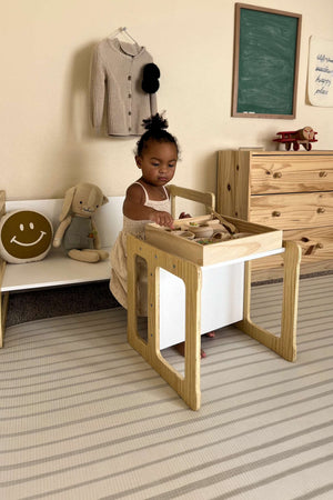 A toddler sits at a wooden convertible table, focused on play. The light-colored furniture blends with a cozy, softly decorated room featuring a chalkboard and plush toys.