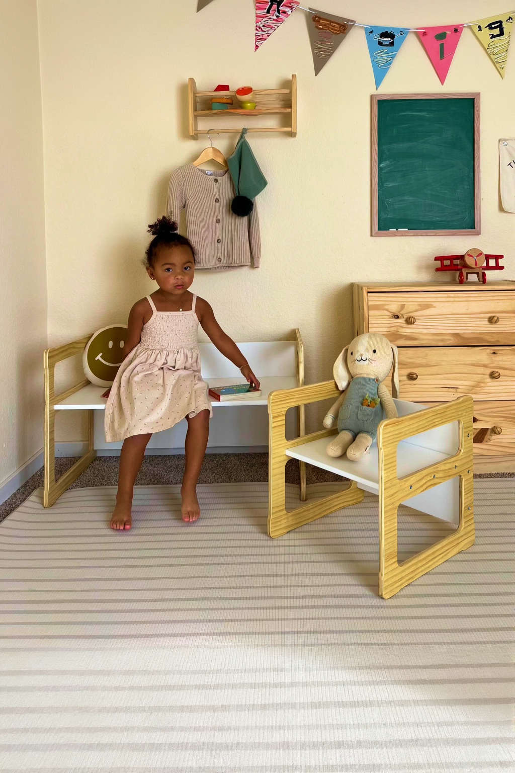 A young girl in a light pink dress stands beside a wooden toddler bench set in a bright room, featuring a chalkboard and decorated with colorful bunting.