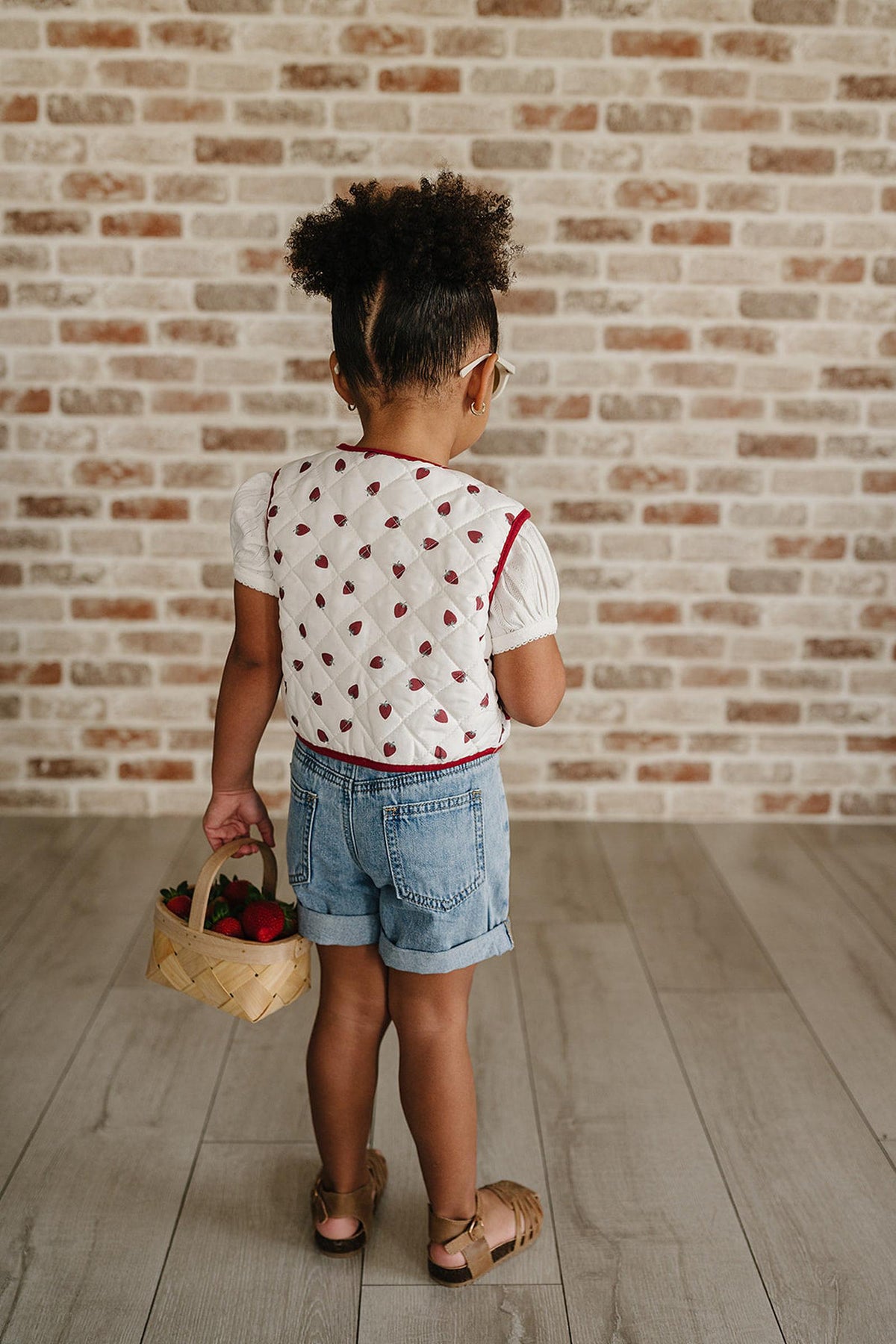 A child stands with their back to the camera, wearing a white quilted vest with a red trim and denim shorts.