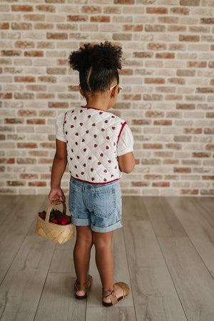 A child stands with their back to the camera, wearing a white quilted vest with a red trim and denim shorts.