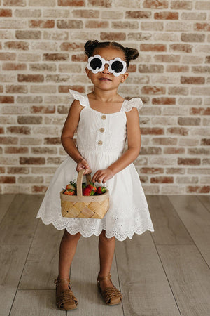 A young girl stands against a brick wall, wearing a white eyelet cotton dress with flutter sleeves and buttons, paired with oversized sunglasses and holding a basket of strawberries.