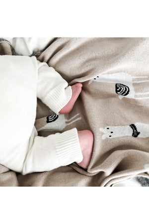 A babys feet rest on a beige knit blanket with llama designs, next to cream-colored clothing.