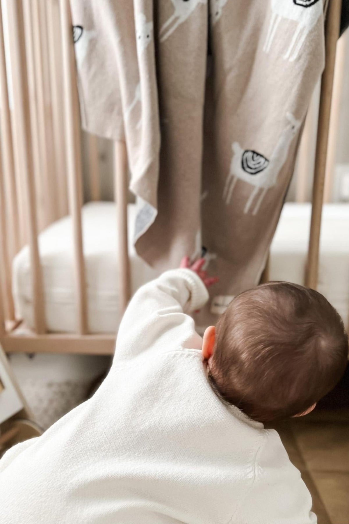 A baby reaches for a beige blanket with llama patterns hanging on a wooden crib.
