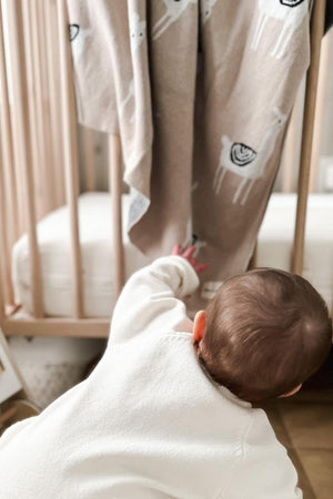 A baby reaches for a beige blanket with llama patterns hanging on a wooden crib.