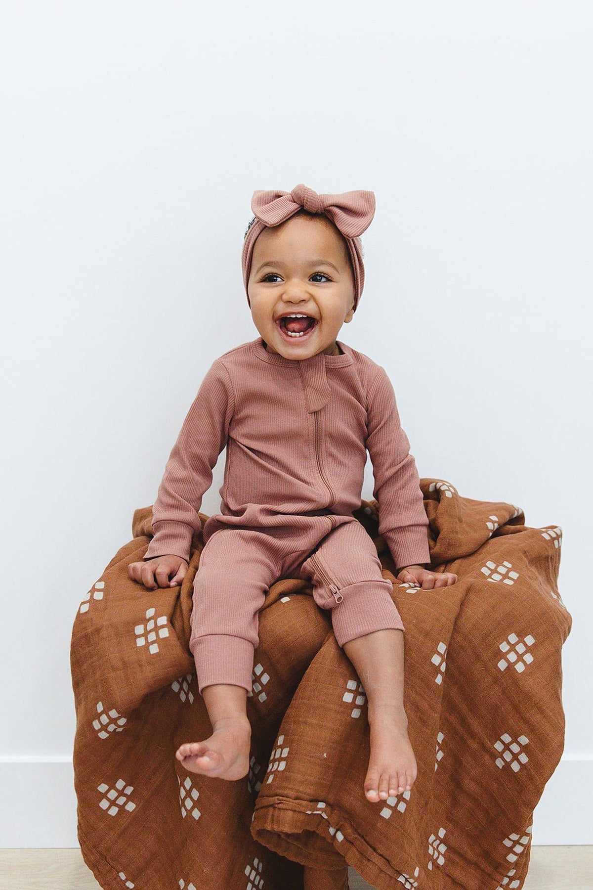 A smiling baby wearing a dusty rose ribbed outfit and head wrap sits on a patterned brown blanket.