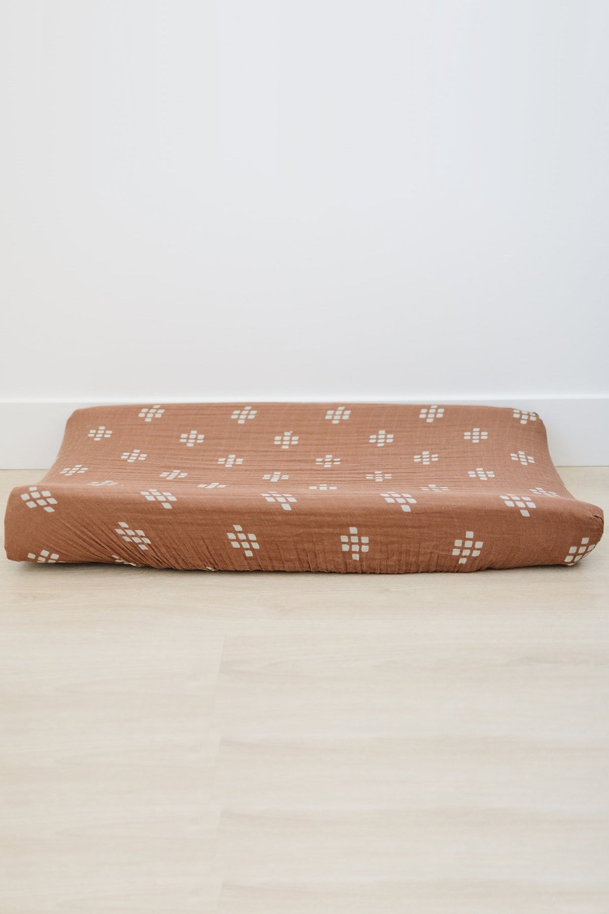 Brown muslin changing pad cover with white geometric patterns on a light wood floor. Made from soft, breathable fabric.