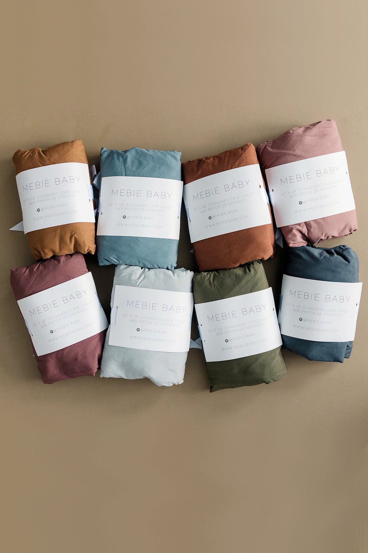 Seven Mebie Baby crib sheets in various colors, neatly folded with labels, displayed on a beige background.