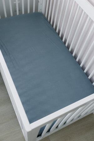 White crib with a dusty blue bamboo stretch sheet, set on a light wood floor. Clean and minimal design.