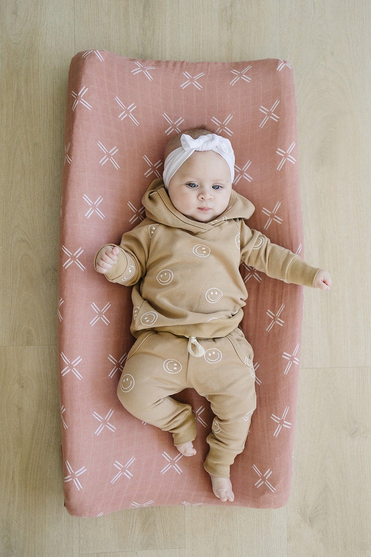 A baby wearing a tan outfit with smiley faces lies on a peach muslin changing pad cover with white patterns.