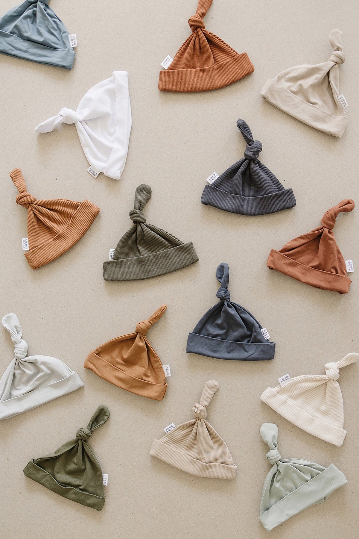 Assorted organic cotton baby knot hats in various neutral colors are displayed on a beige background.