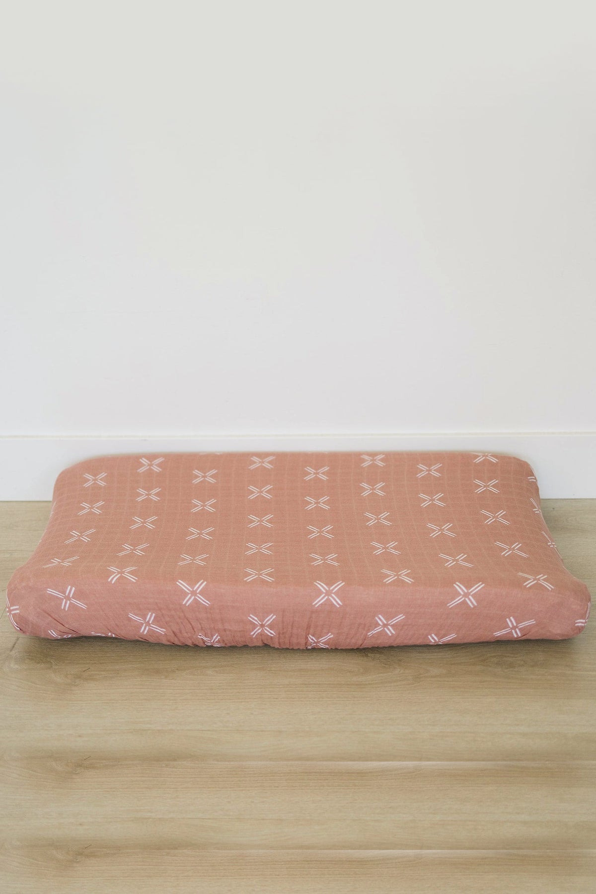 Peach-colored muslin changing pad cover with white cross patterns, placed on a wooden floor against a plain wall.