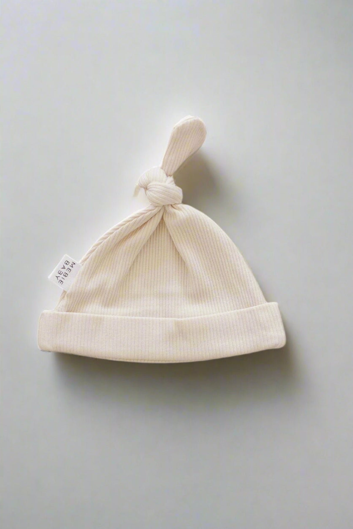 Cream-colored ribbed newborn knot hat made from organic cotton with a small brand tag, set on a plain background.