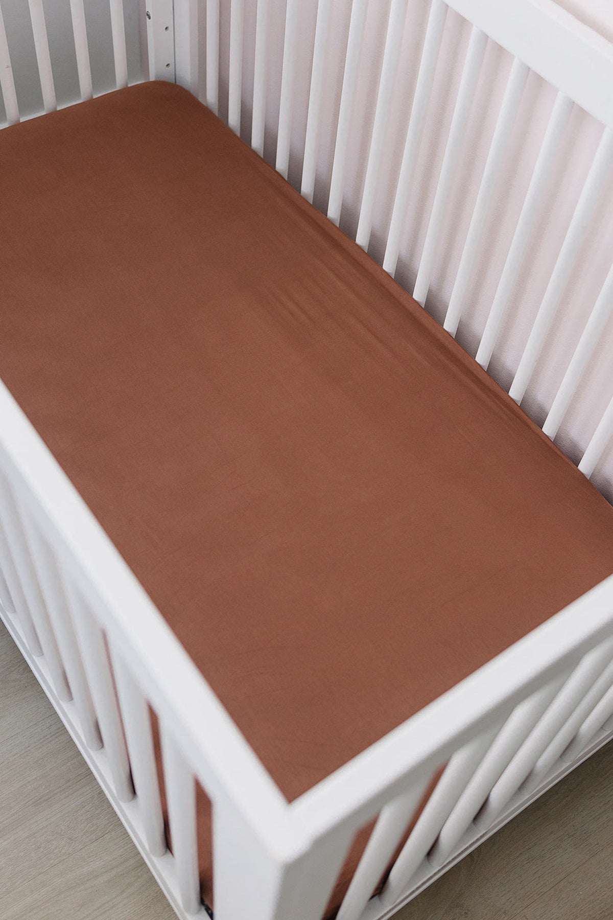 White crib with a rust-colored crib sheet, featuring a smooth, soft bamboo fabric. Set on a wooden floor.