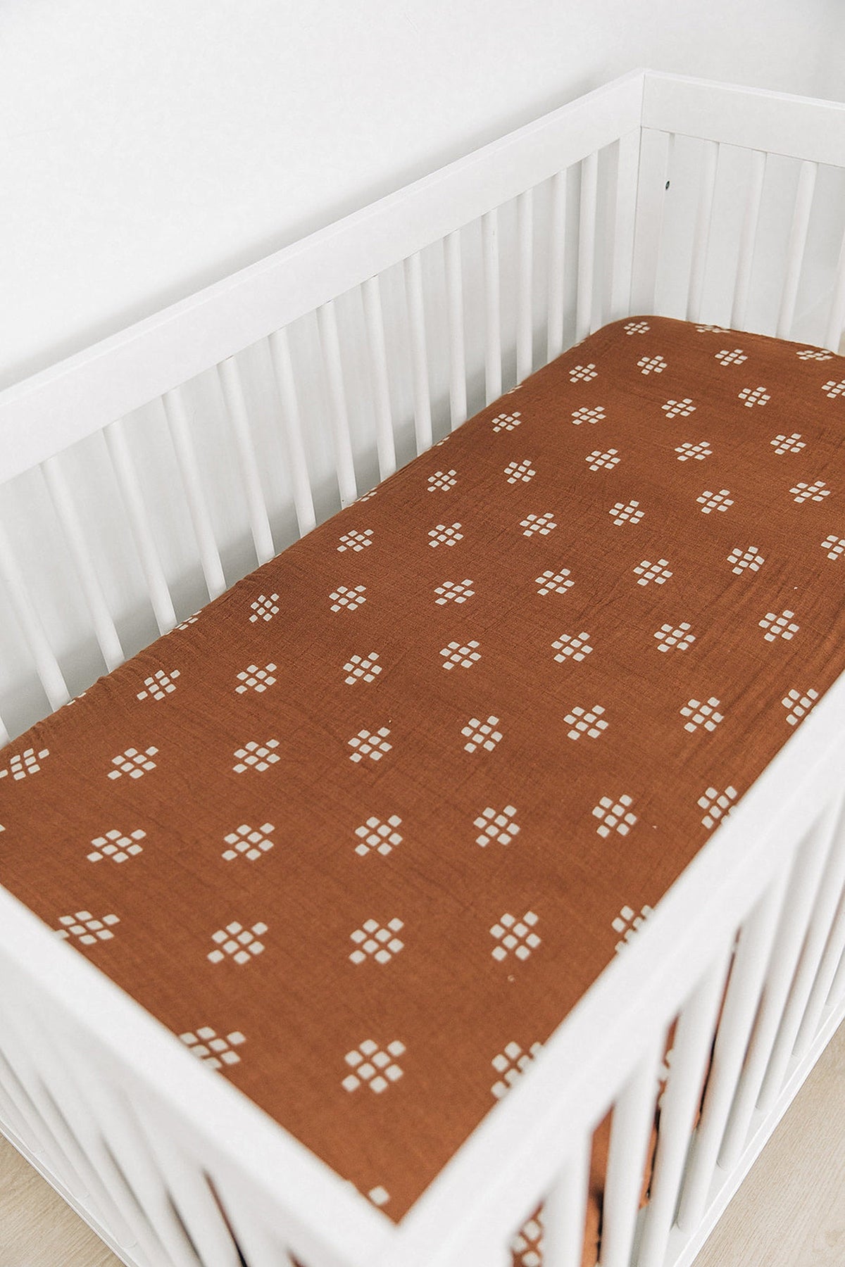 White crib with a chestnut muslin sheet featuring a geometric pattern, highlighting a cozy and stylish nursery setting.