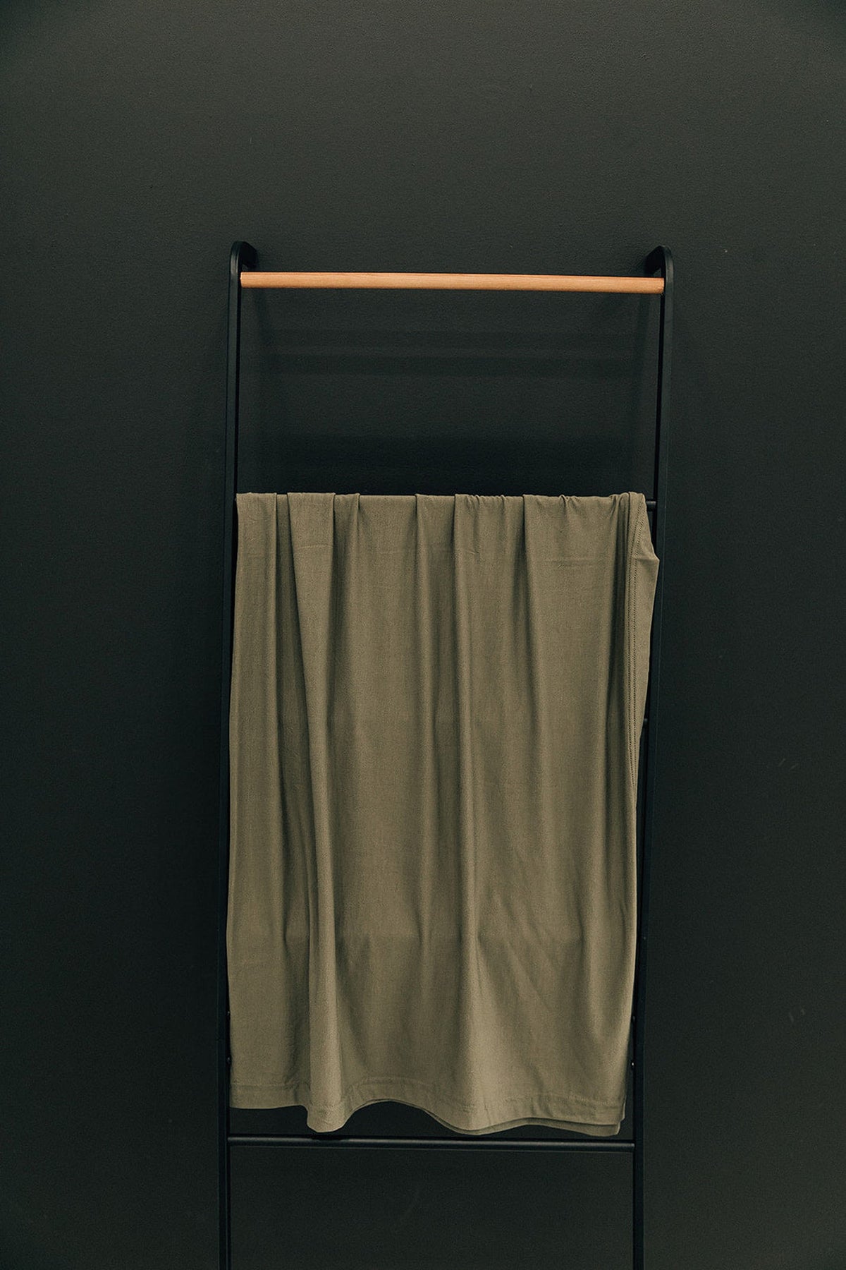 An olive green swaddle blanket made of bamboo fabric hangs on a black metal and wooden rack.