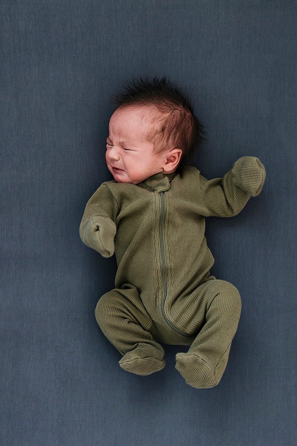 Newborn in olive green onesie on a charcoal crib sheet, expressing discomfort with eyes closed.