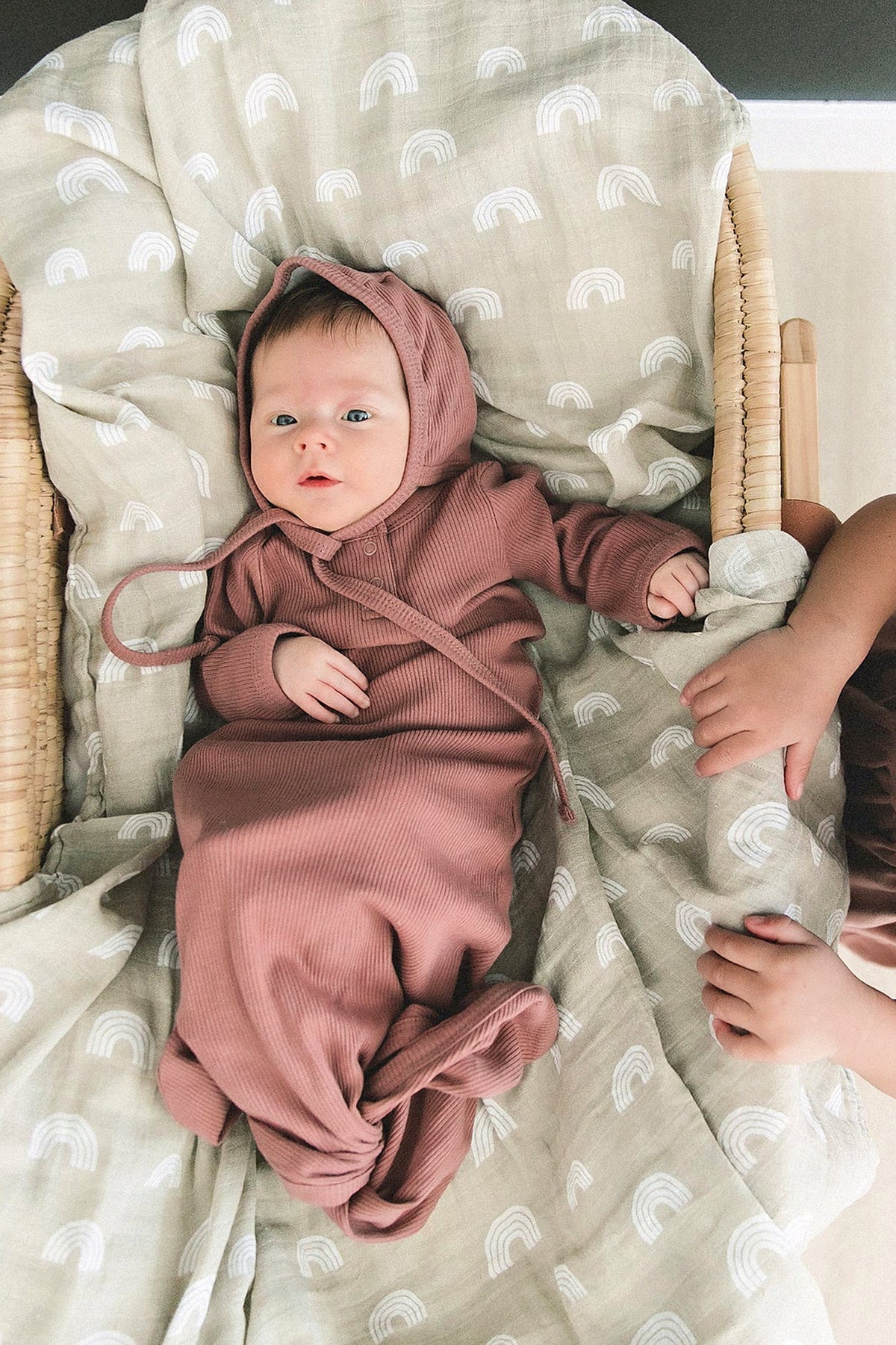A baby wearing a dusty rose ribbed knot gown lies on a patterned blanket in a wicker bassinet.