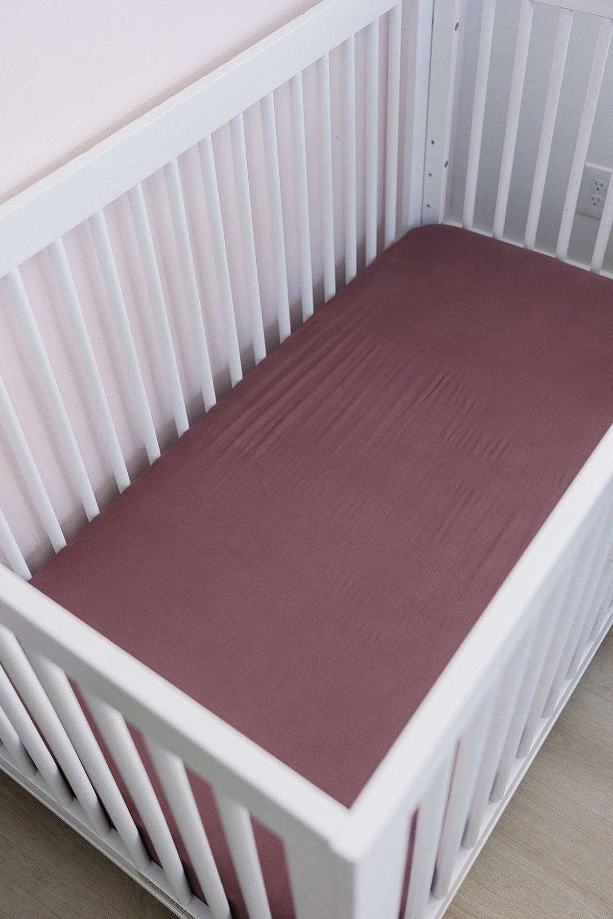 White crib with a plum-colored bamboo stretch sheet, set against a light pink wall.