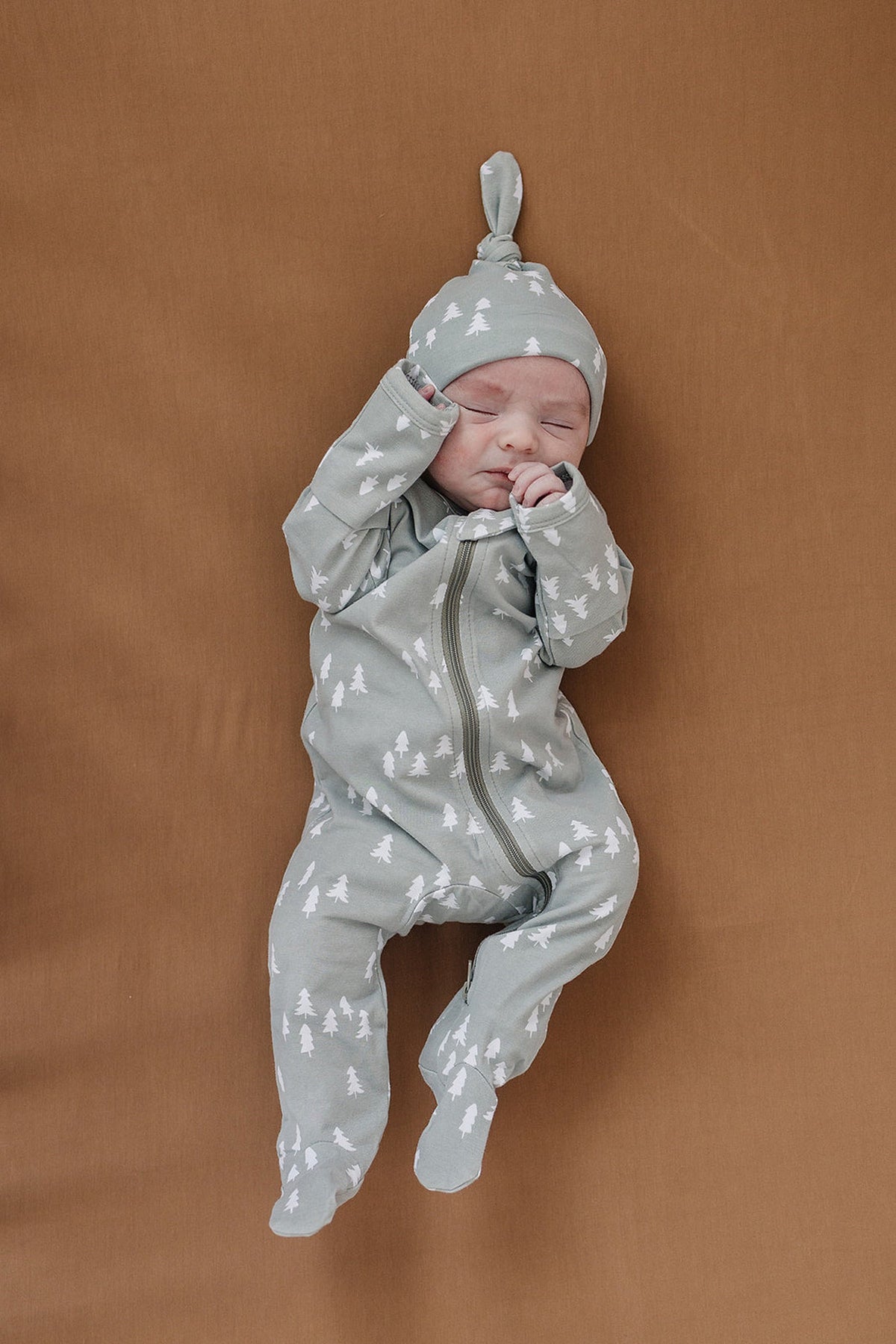 A baby in a tree-patterned, sage green onesie and matching hat lies on a mustard-colored surface.