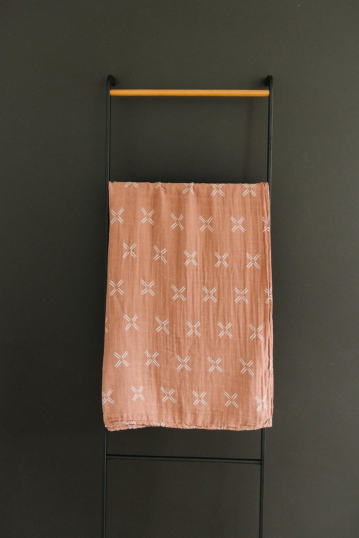 A peach-colored muslin swaddle blanket with white cross patterns hangs on a black metal rack.