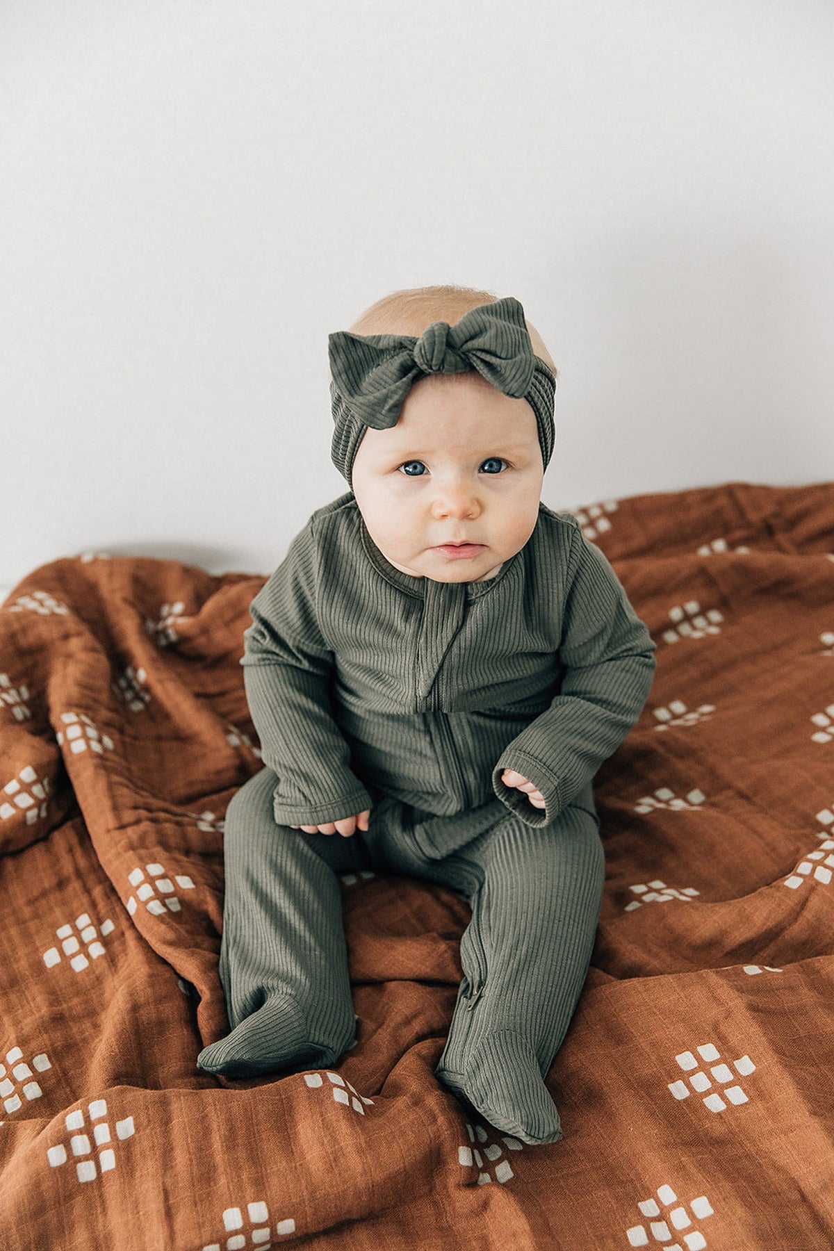 A baby wearing a green ribbed outfit and matching headband sits on a patterned brown muslin quilt.