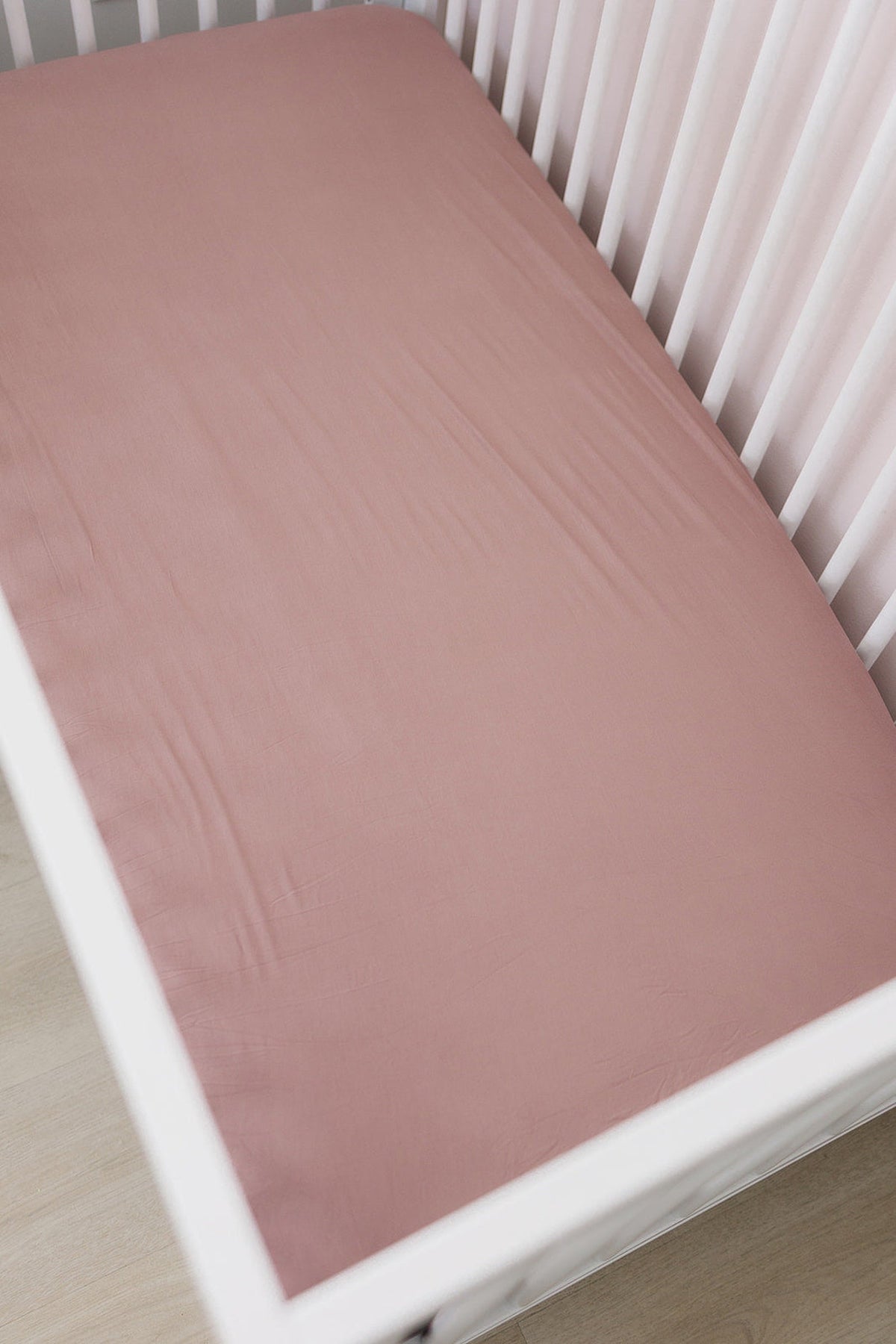 Dusty rose crib sheet made from bamboo fabric, fitted over a white crib in a softly lit room.