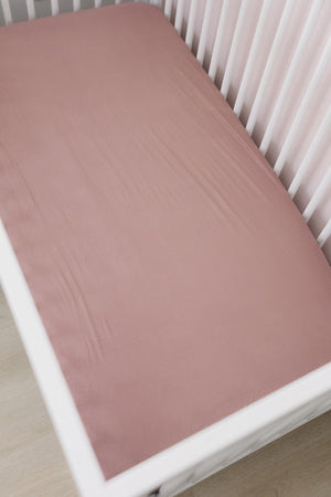 Dusty rose crib sheet made from bamboo fabric, fitted over a white crib in a softly lit room.