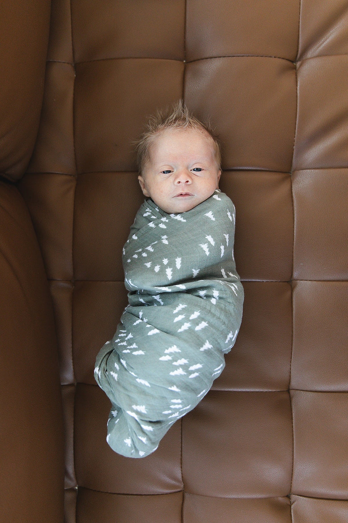 A baby wrapped in a green muslin swaddle with white pine patterns, lying on a brown cushioned surface.