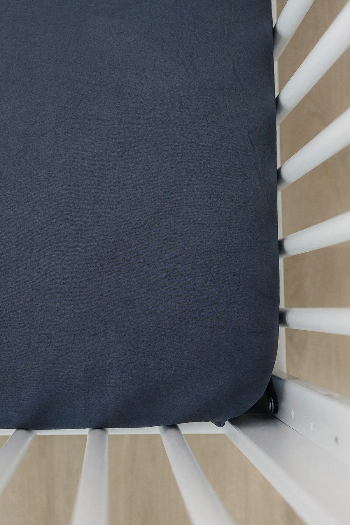 Dark charcoal crib sheet on a white crib with wooden flooring visible beneath. Fabric appears soft and smooth.