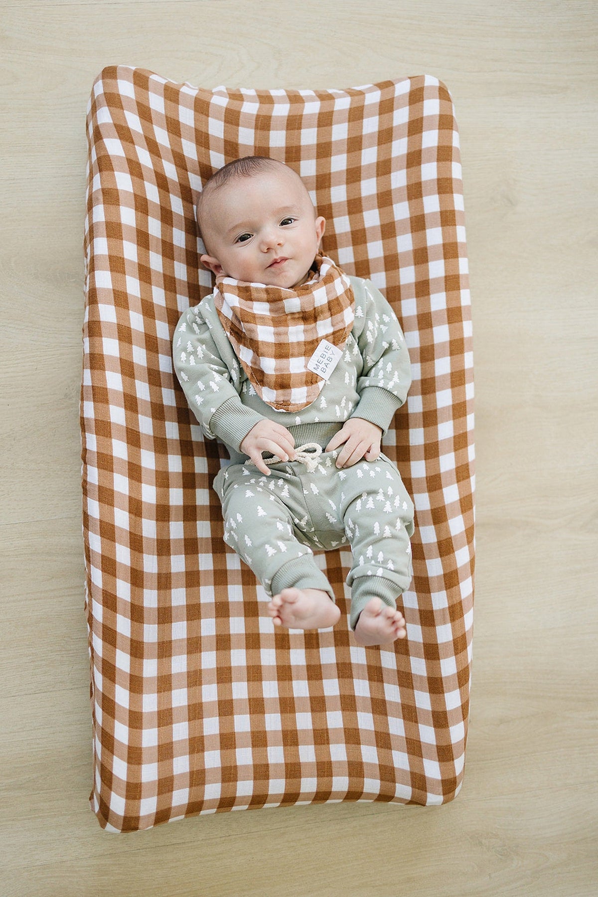 A baby wearing a brown and white gingham bib lies on a matching muslin changing pad cover.