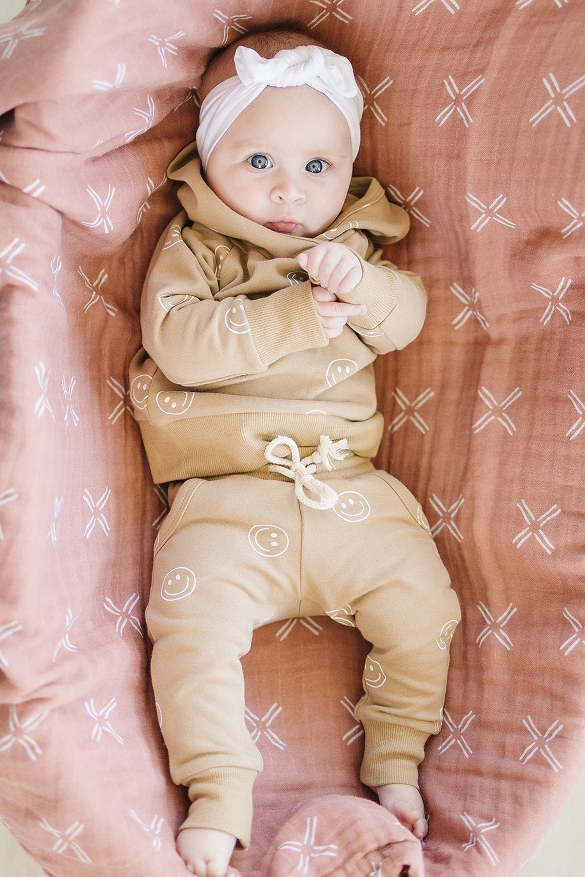 A baby lies on a peach muslin quilt, wearing a tan outfit with smiley faces and a white headband.