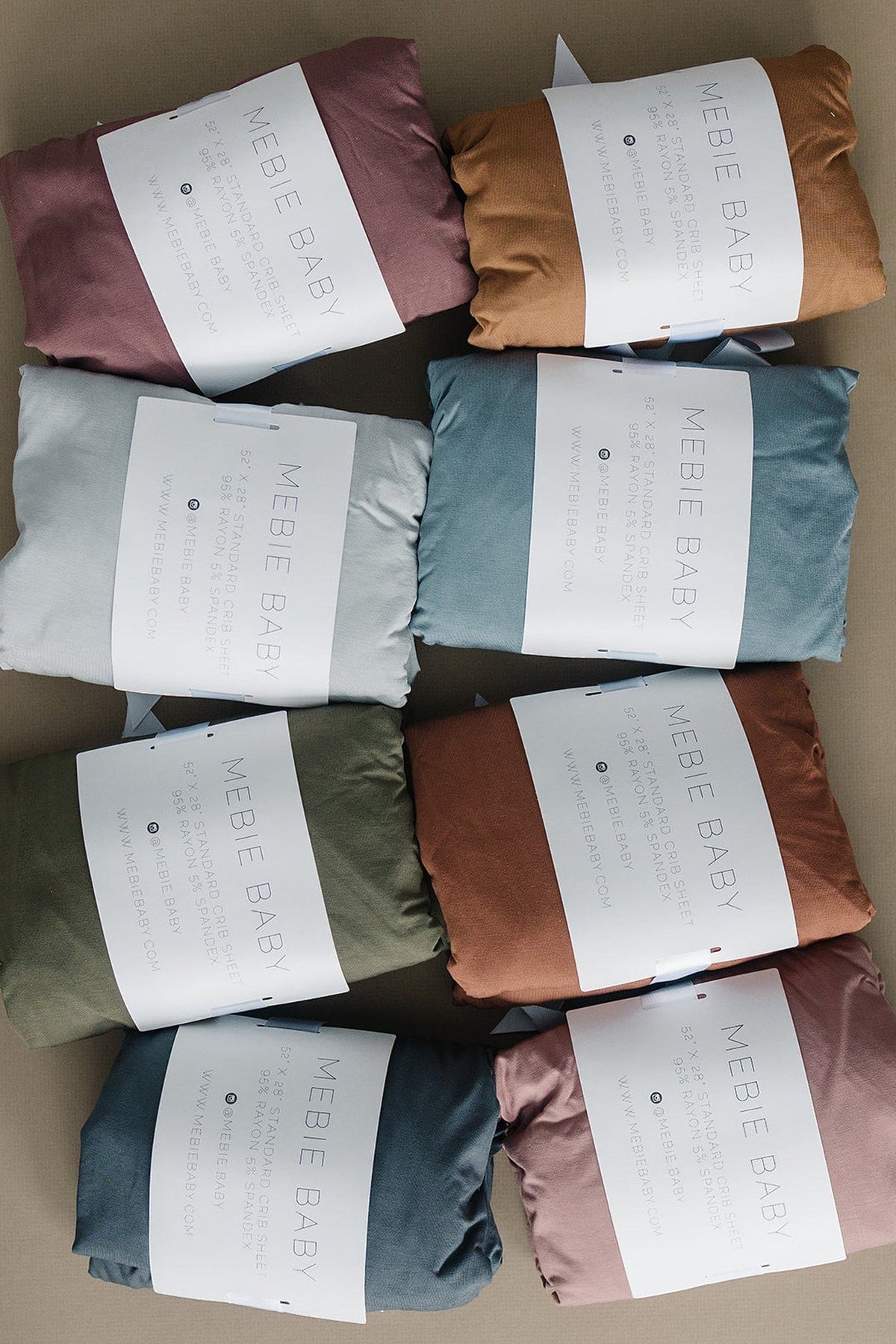 Assorted Mebie Baby crib sheets in various colors are neatly folded with branded labels on each package.