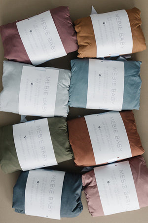 Assorted Mebie Baby crib sheets in various colors are neatly folded with branded labels on each package.