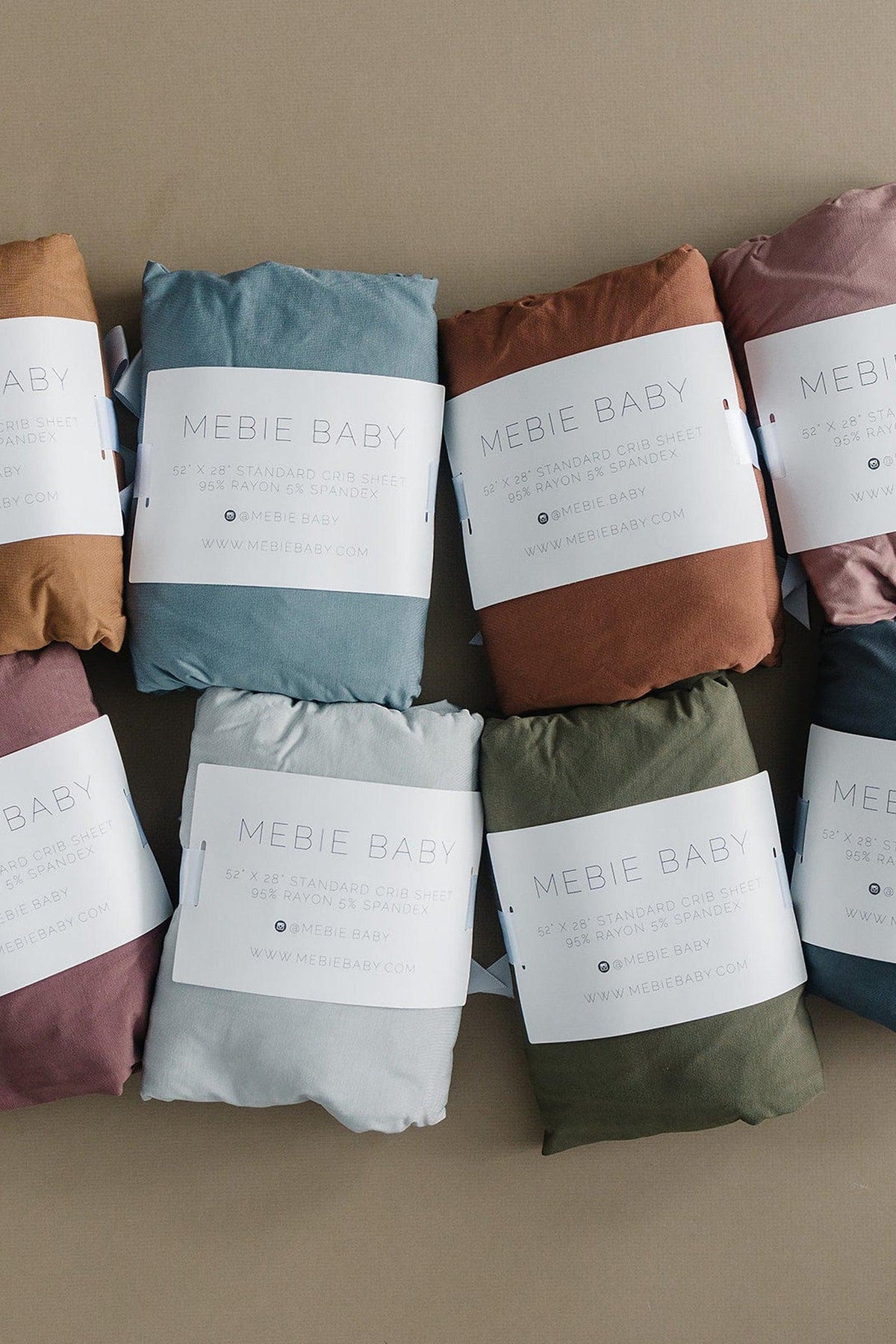 Various colored Mebie Baby crib sheets are displayed, wrapped in labels. They feature soft, eco-friendly fabric.
