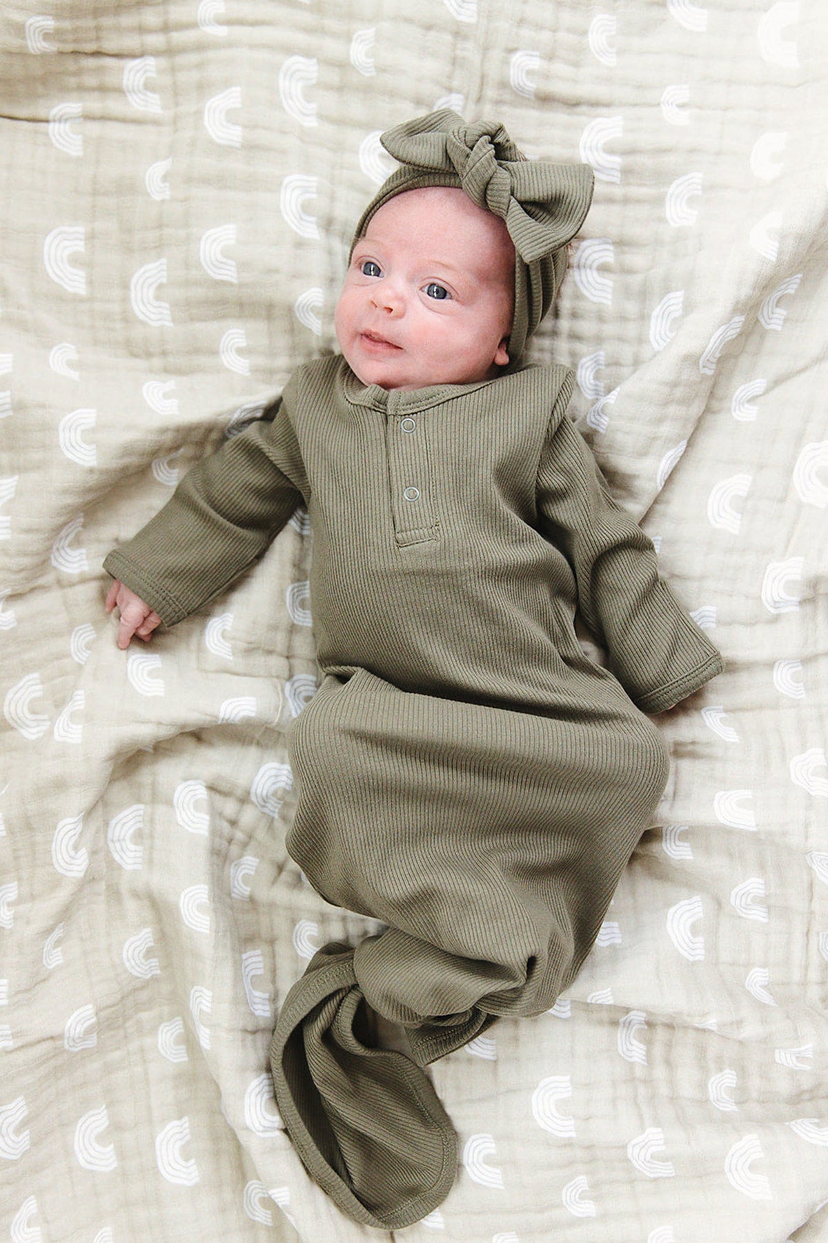 Baby in olive ribbed outfit and head wrap on a patterned beige blanket, smiling gently.