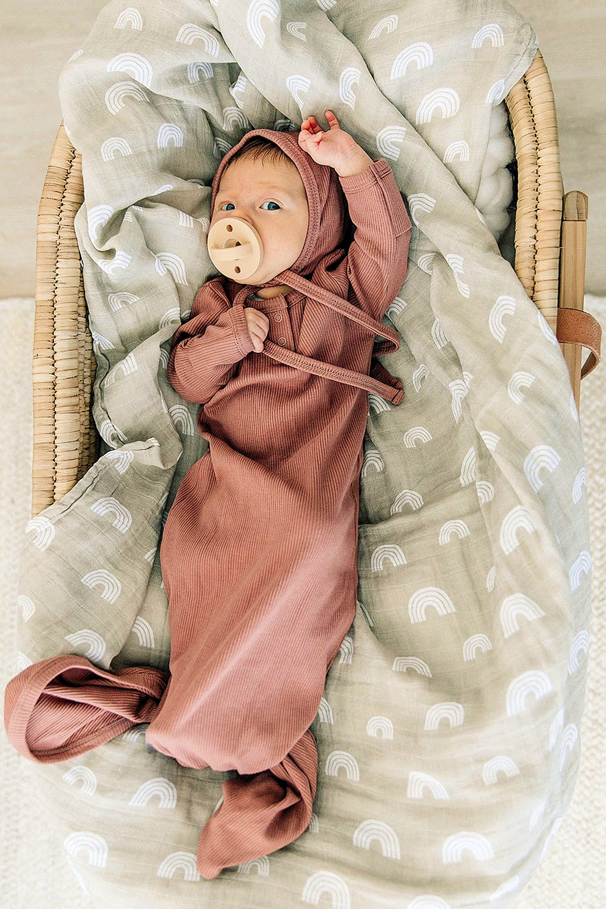Newborn in a dusty rose ribbed knot gown with a pacifier, lying on a rainbow-patterned blanket.