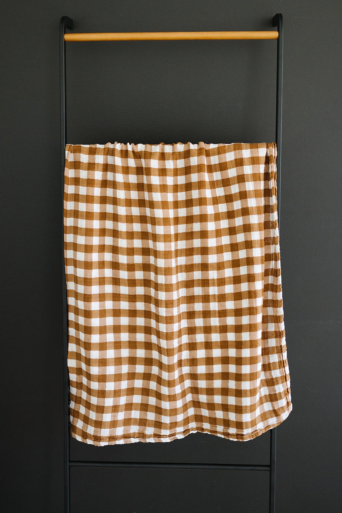 Brown and white gingham muslin swaddle blanket hanging on a black metal and wood rack.