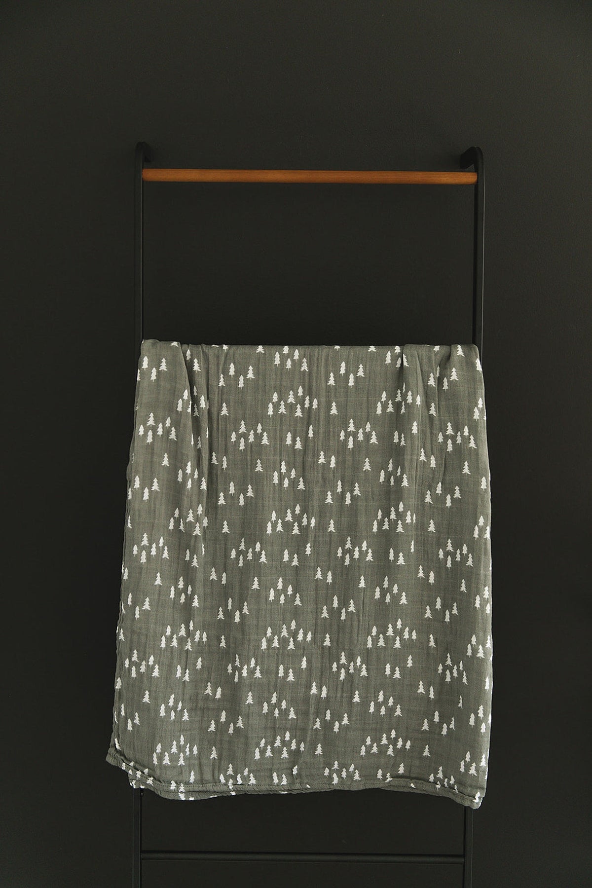 Gray muslin swaddle blanket with white tree patterns hanging on a black and wooden rack.