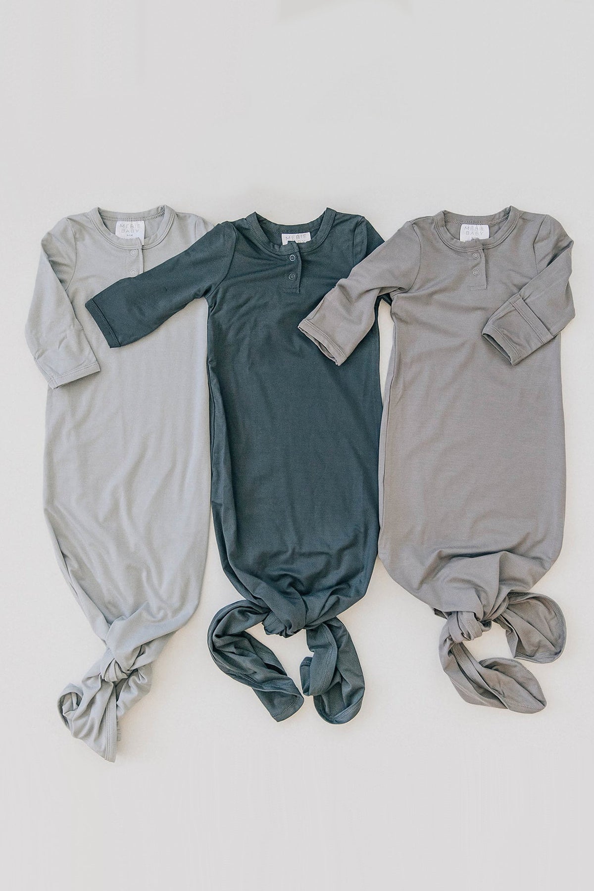 Three baby knot gowns in light gray, charcoal, and taupe are laid out side by side on a light background.