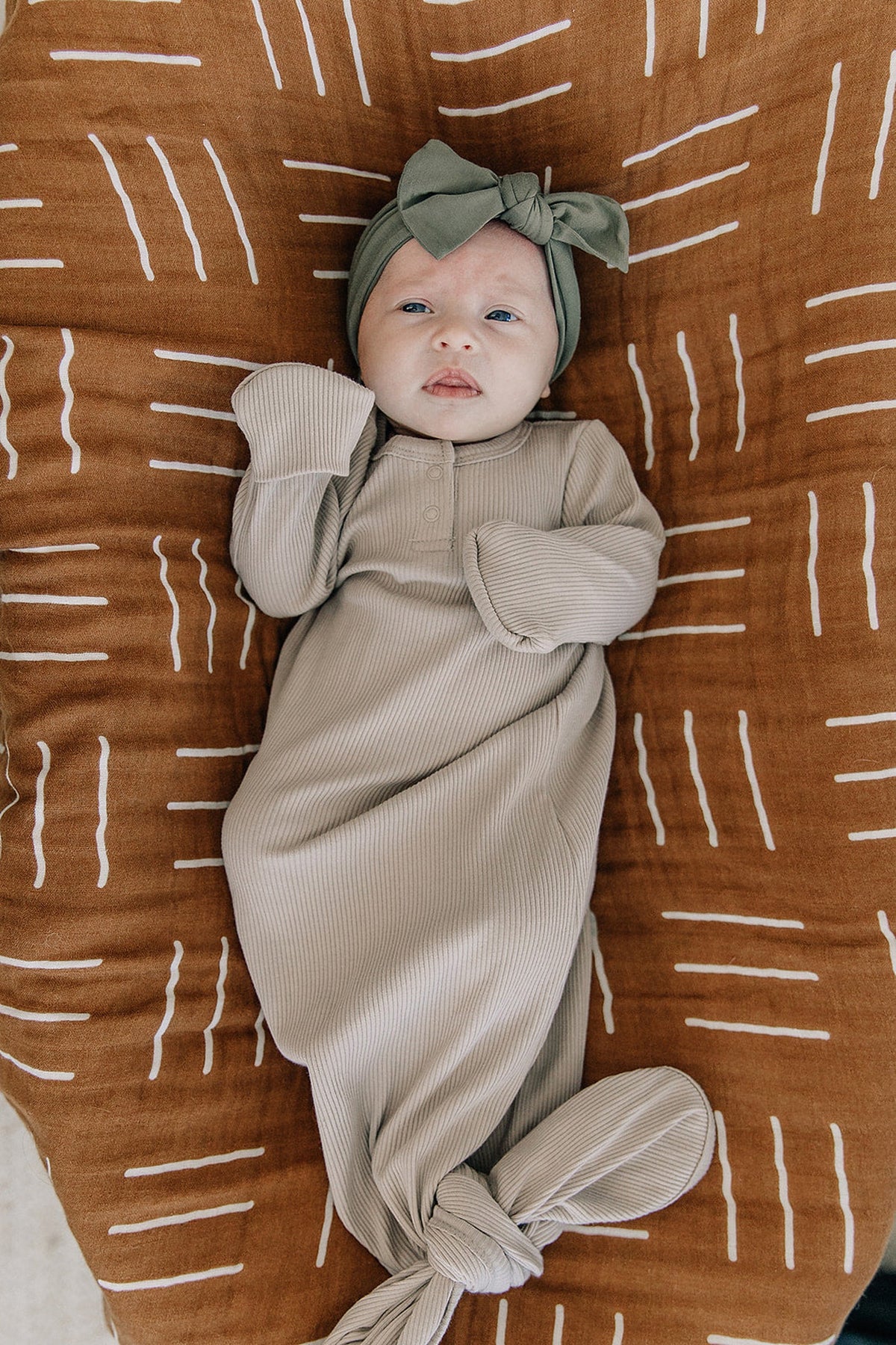 A baby lies in an oatmeal ribbed gown with a knotted bottom and a green headband on a patterned blanket.