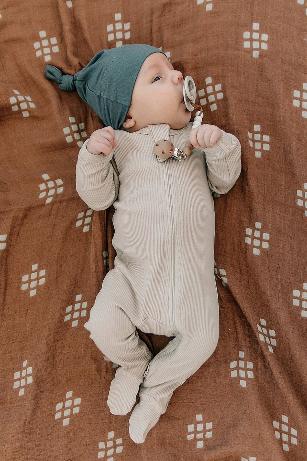 A baby lies on a patterned brown blanket wearing oatmeal-colored ribbed pajamas and a green hat, holding a pacifier.